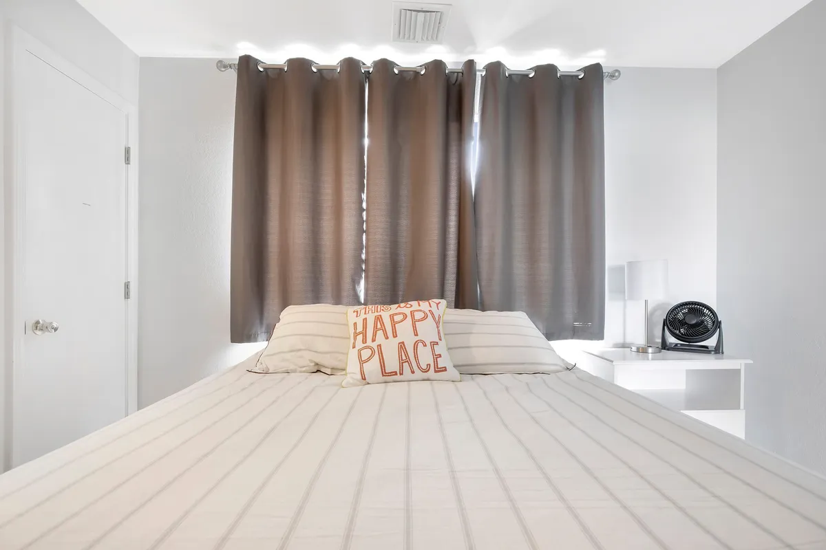 A comfortable bedroom featuring a plush bed with a decorative pillow, soft curtains framing a window, and a nightstand with a lamp. The setting is serene and welcoming, bathed in natural light, ideal for restful downtime or afternoon naps.
