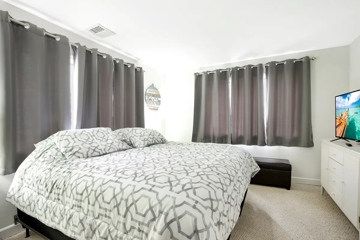 A cozy bedroom with a large bed dressed in patterned bedding, gray curtains framing a window, and a television atop a dresser. The room is softly lit, offering a peaceful oasis for winding down and enjoying quiet privacy.