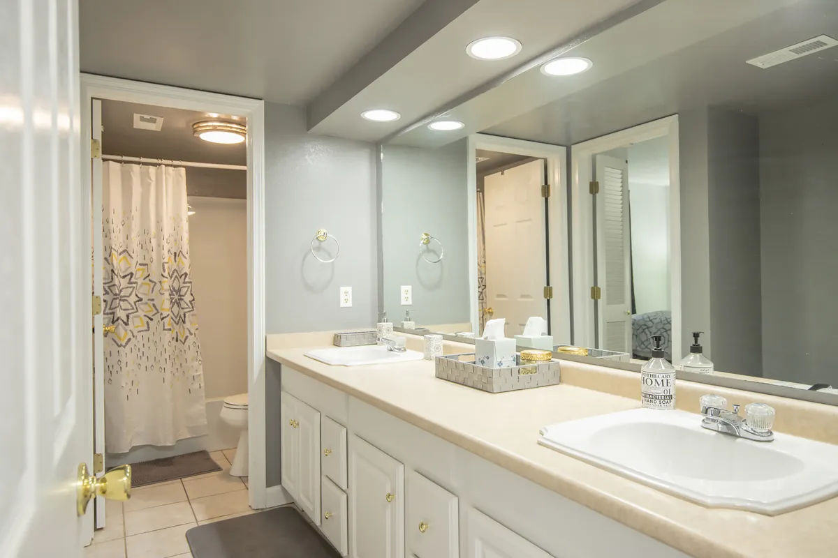 A fresh, inviting bathroom with double vanity sinks, a mirrored cabinet, and a glimpse of a modern shower area. The overall décor is clean and serene, suggesting a spa-like atmosphere for morning routines or evening relaxation.