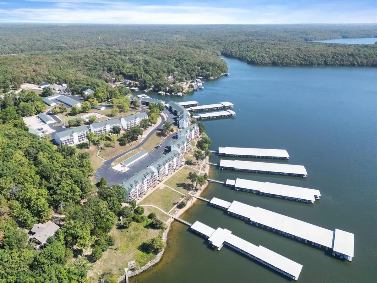 An aerial or elevated view of the expansive lake, marina, surrounding forest, and nearby buildings. The scene captures the scale of the Lake of the Ozarks, highlighting both natural beauty and recreational opportunities on the water.