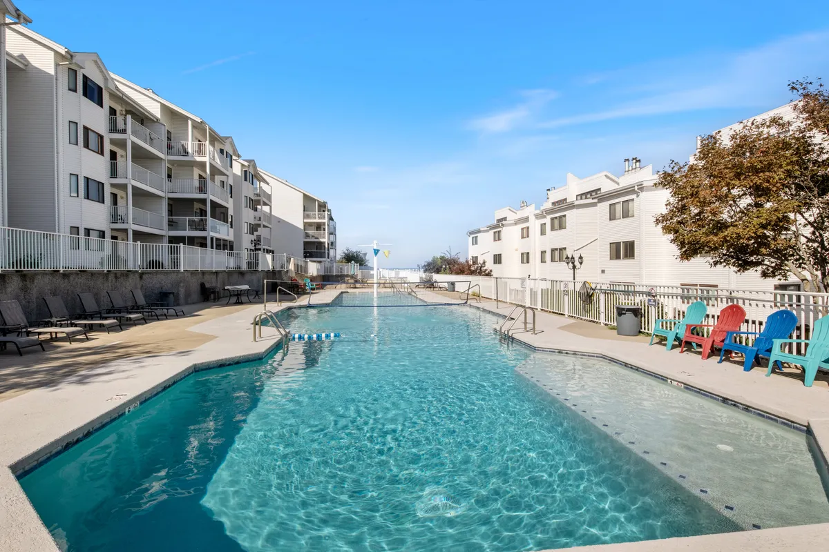 The hero image showcases a sparkling resort-style swimming pool framed by vibrant Adirondack chairs, with the condo buildings and lush landscaping in the background. The pool area is the focal point, inviting guests to relax, swim, and socialize in a sun-drenched, leisurely setting. The image hints at sunset lighting, enhancing the inviting atmosphere and lakeside vibe.