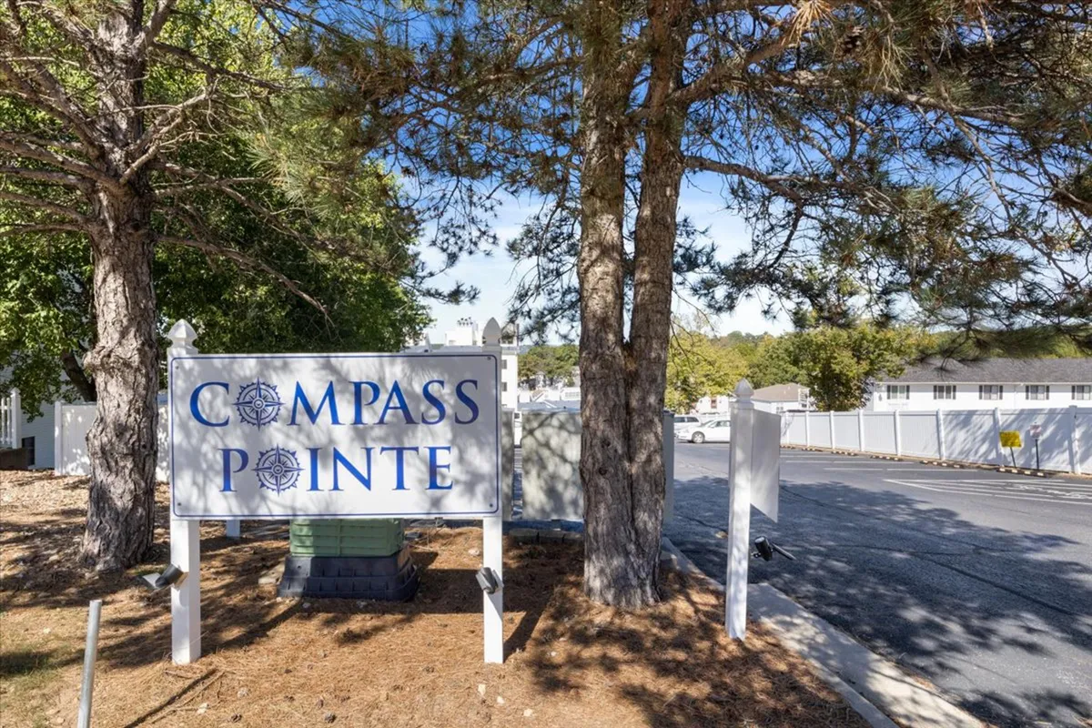 A wide shot of the Compass Pointe sign positioned amidst lush green trees with a well-paved roadway leading into the community. The entrance feels open and welcoming, hinting at a well-maintained, accessible neighborhood.