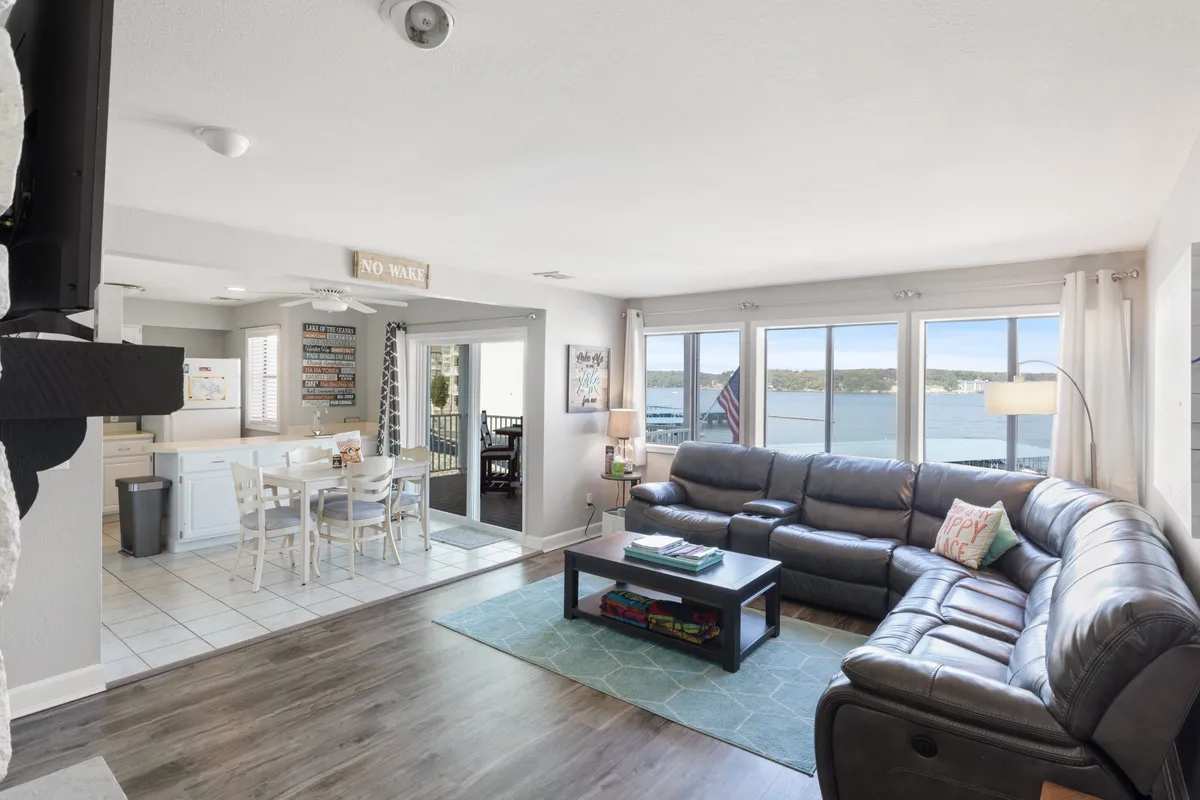A welcoming, sunlit living room featuring large windows overlooking the water. The space is furnished with a comfortable seating area, a dining table, and charming decorative touches, including a nautical 'NO WAKE' sign. The overall atmosphere is casual and inviting, with ample light and spectacular lake views.