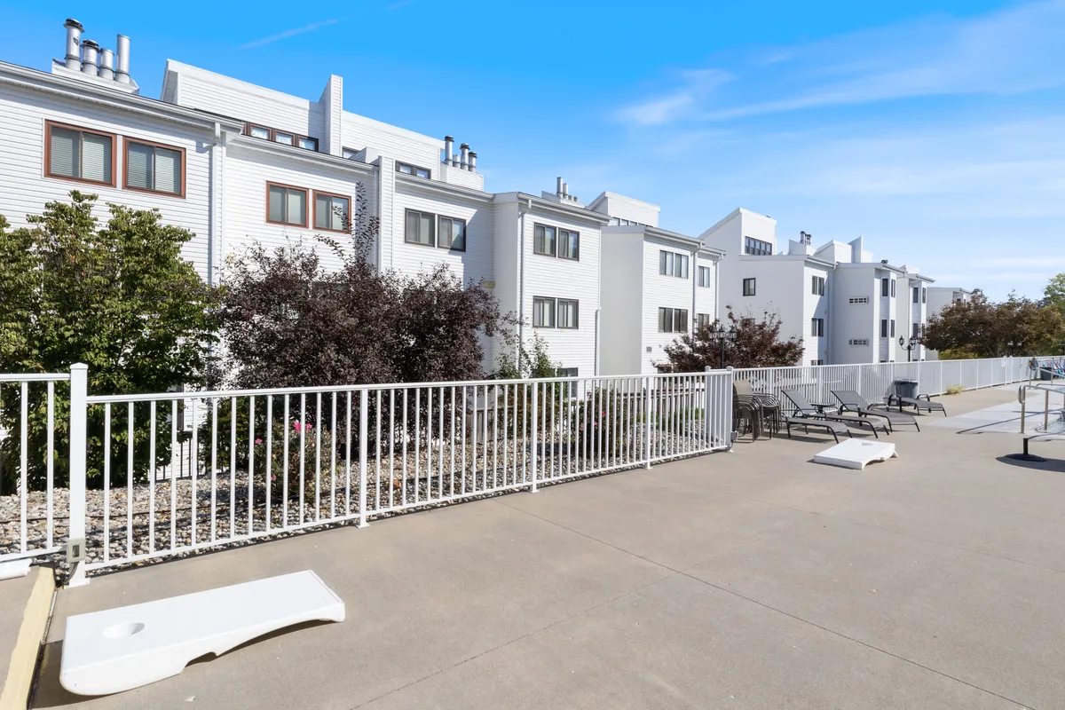 Modern white condo buildings surrounded by lush green shrubbery and a fenced patio area with lounge chairs. The scene captures a relaxed, contemporary environment perfect for group socializing, sunbathing, or enjoying coffee outdoors.