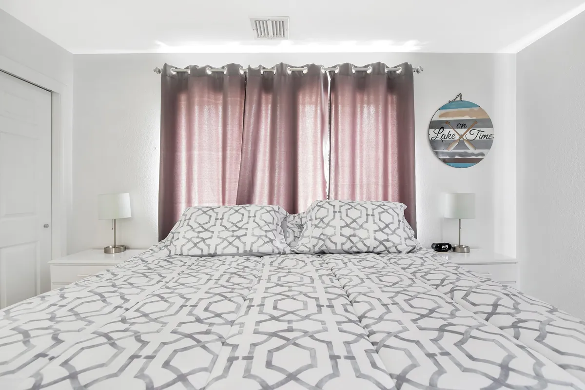 A tranquil bedroom featuring a bed with geometric patterned bedding, soft curtains filtering gentle sunlight, and a decorative wall sign above the headboard. The space feels airy and calm, inviting relaxation and restful sleep.