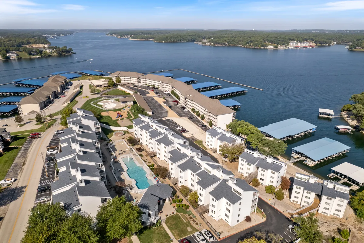 This exterior image captures a waterfront view with a large swimming pool in the foreground, boat docks extending into the lake, a playground area, and landscaped gardens. The scene is vibrant and inviting, showcasing multiple resort-style amenities and the scenic lake backdrop.