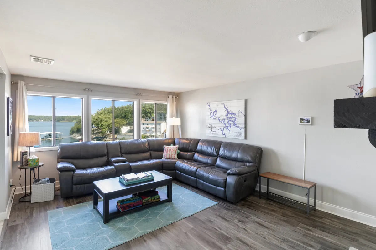 A spacious and cozy living area with a large sectional sofa and coffee table, set before a wall of windows offering panoramic lake views. The space is styled for both comfort and group interaction, ideal for relaxing or entertaining guests while enjoying the scenic backdrop.