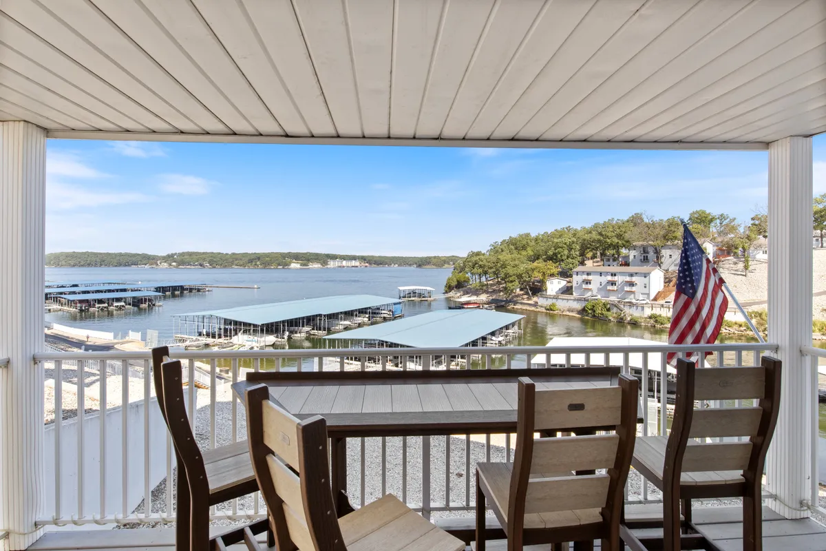 A tranquil outdoor seating area with a clear view of the lake, boathouses, and an American flag waving gently. The scene is set for relaxing after a day on the water, enjoying a coffee in the morning or a sunset in the evening. Boats are visible in the distance, reinforcing the connection to water recreation.
